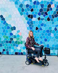 Amy, a disabled, white Australian woman with blonde hair, sits on her mobility scooter in front of a wall with blue and white geometric shapes.